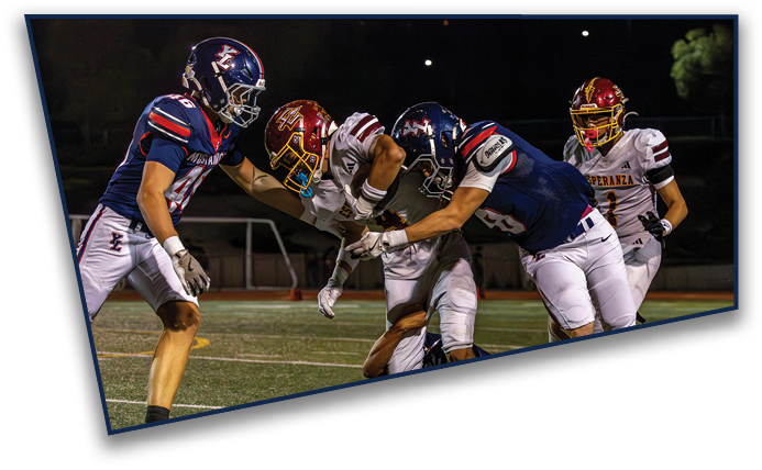 Four people in football uniforms are actively engaged in a nighttime game on a field, with two players in dark blue and two in white and red helmets. Description generated by AI