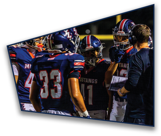 People wearing football helmets and uniforms are gathered closely while one person wearing a headset holds a clipboard and appears to be giving instructions. Description generated by AI