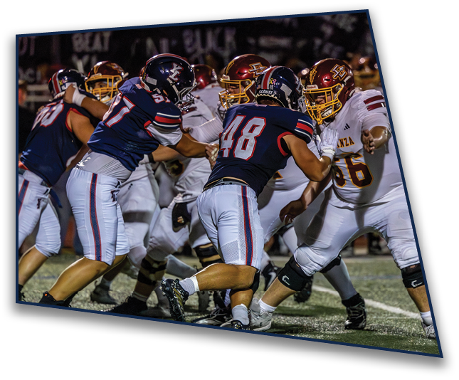 Several football players in dark and white uniforms are engaged in a close, intense play on a well-lit field during a game. Description generated by AI
