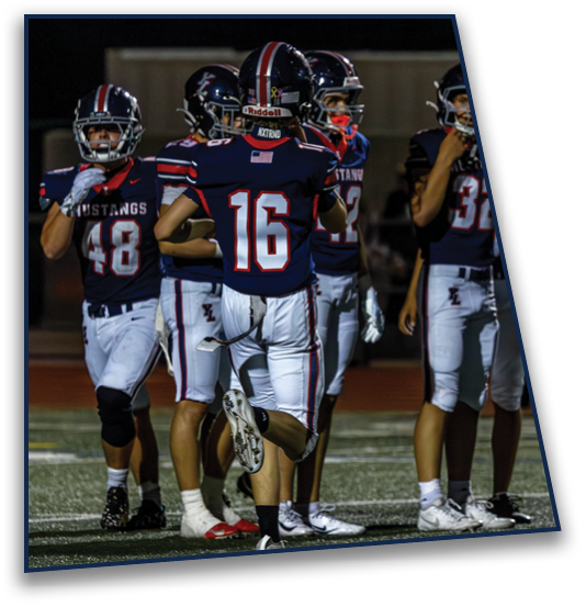 Five people wearing dark blue football uniforms with white pants and helmets stand on a field at night, with one person running toward the group. Description generated by AI