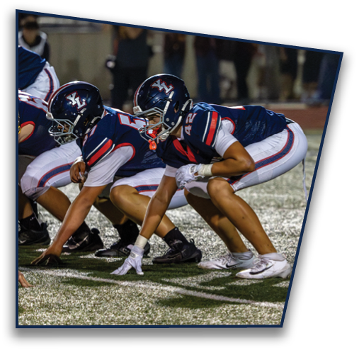 Two people in football uniforms and helmets are crouched in a ready position on a turf field during a game. Description generated by AI