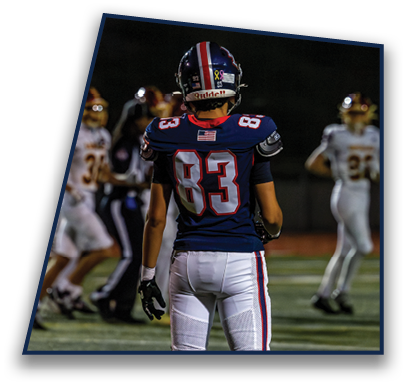 A person wearing a dark blue football uniform with the number 83 on the back stands on a field facing away from the camera while other players and a referee are visible in the background. Description generated by AI