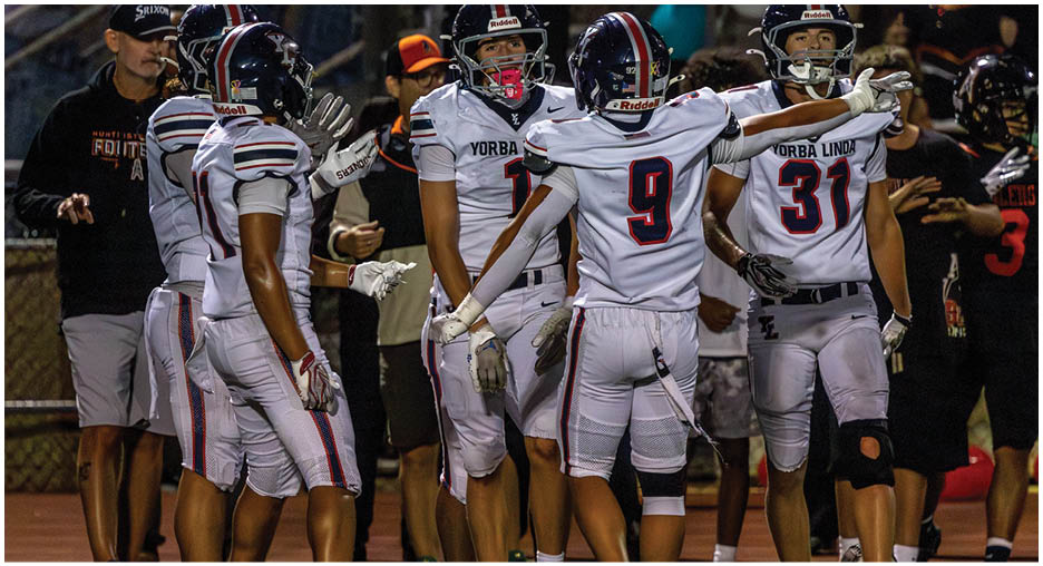A group of football players in white uniforms with black helmets are gathered on a field with one player extending arms outward while others stand nearby. Description generated by AI