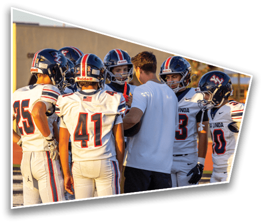 People wearing sports uniforms are standing together on a field with one person in shorts and a t-shirt among them. Description generated by AI