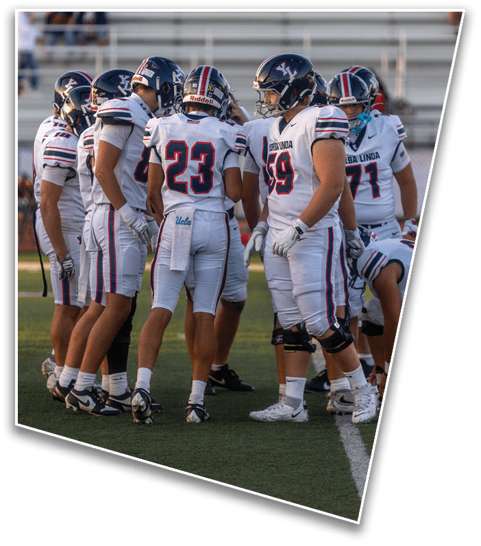 A group of people wearing white football uniforms with helmets are huddled together on a field during a game or practice. Description generated by AI