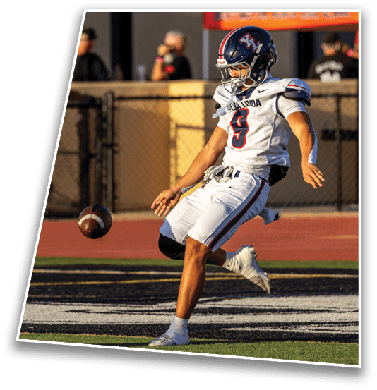 A person in a football uniform is kicking a football on a field near the end zone during a game. Description generated by AI