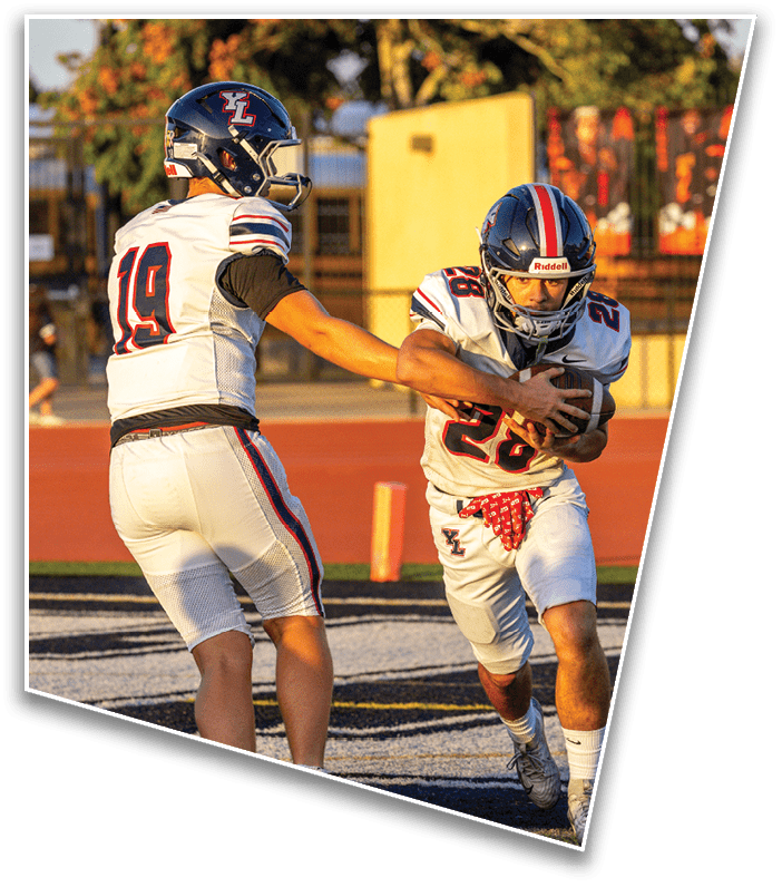 Two people in football uniforms are on a field with one handing off the ball to the other during a game. Description generated by AI
