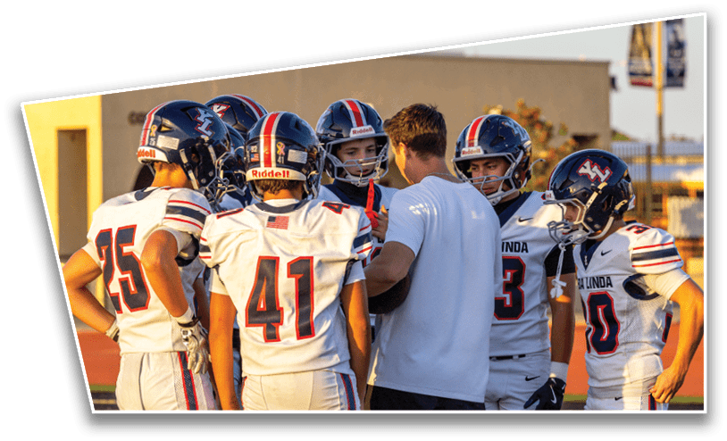 A group of people wearing football uniforms and helmets are gathered around a person in a white shirt on a sports field during daylight. Description generated by AI