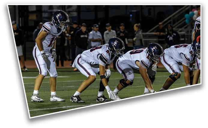 Four people in football uniforms and helmets are positioned on a field, preparing for a play during a game. Description generated by AI