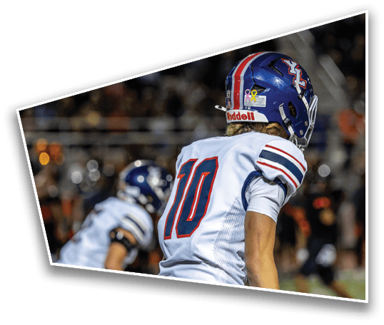 Two people wearing football helmets and white jerseys with red and blue stripes are on a field during a game. Description generated by AI