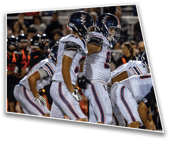 Two people wearing football helmets and white jerseys are on a field with blurred spectators in the background. Description generated by AI