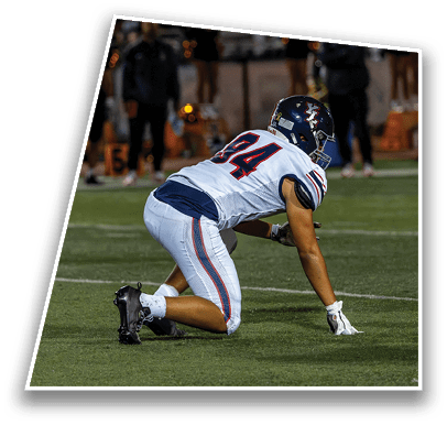 A person in a white sports uniform is kneeling on a football field with one hand touching the ground. Description generated by AI