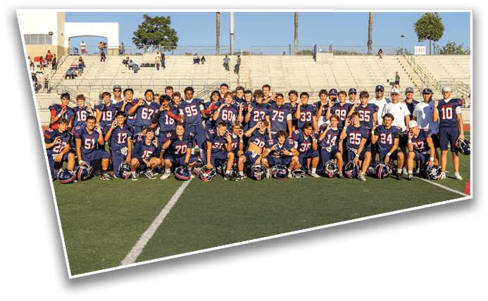 A large group of people wearing matching dark blue football uniforms pose together on a football field with empty bleachers in the background. Description generated by AI