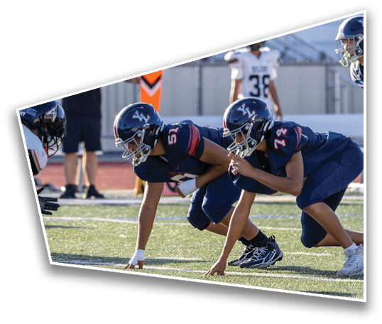 People in football uniforms are lined up on a field with a referee or coach standing near an orange down marker in the background. Description generated by AI