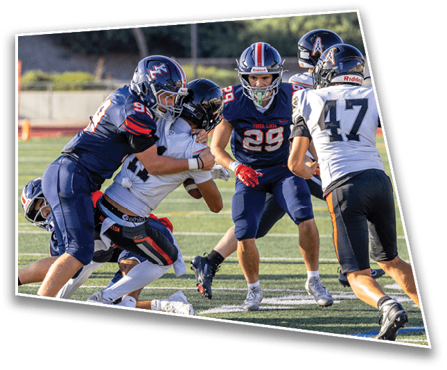 Two people in dark blue football uniforms tackle a person in a white uniform on a football field while another player in a dark blue uniform watches nearby. Description generated by AI