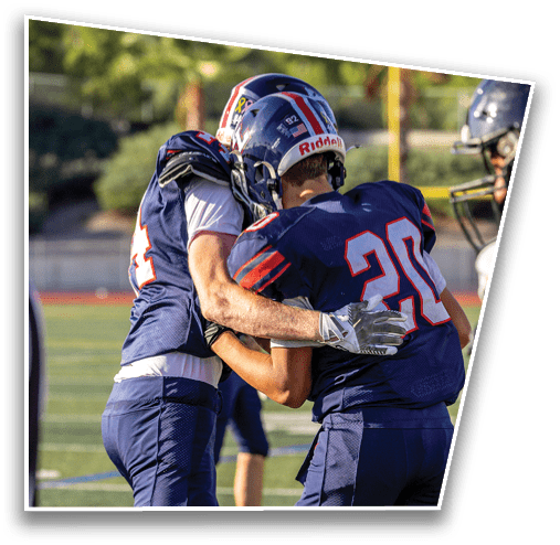 Two people in football uniforms and helmets are embracing on a football field during a game or practice. Description generated by AI