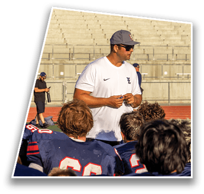 A person wearing a white shirt and cap is speaking to a group of people in blue sports uniforms on a field with empty bleachers in the background. Description generated by AI