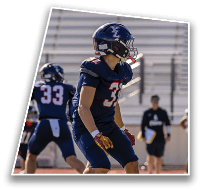 Two people in football uniforms are on a field with one person in the foreground wearing gloves and a number 33 jersey while another person stands in the background holding a clipboard. Description generated by AI