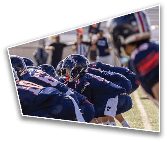 People in sports uniforms are crouched in a ready position on a grassy field, likely preparing for a play in a football game. Description generated by AI