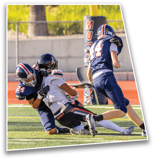 Two people in football uniforms are engaged in a tackle on a grassy field with a track in the background. Description generated by AI