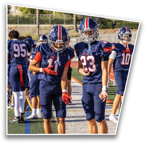 Two people in football uniforms stand on a field with one looking down at their hands and the other looking forward while other players are in the background. Description generated by AI