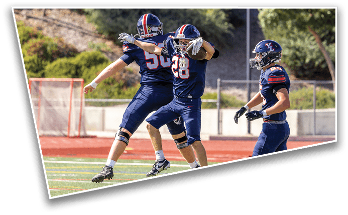 Three people in football uniforms are on a field with one person celebrating with arms outstretched while the others look on. Description generated by AI