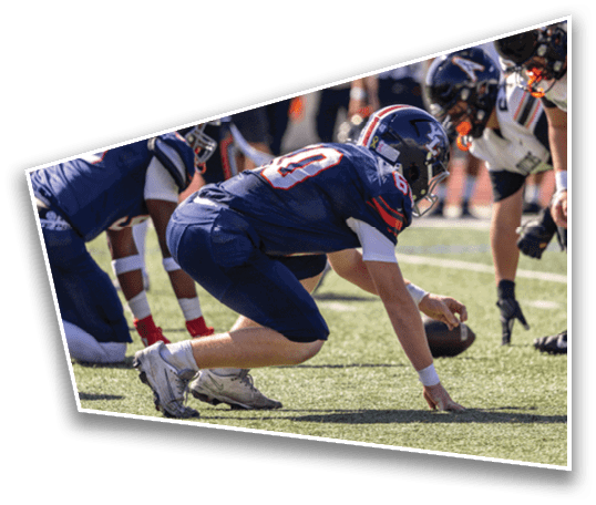 A person in a blue football uniform is crouched on the field holding a football while facing a group of opposing players in white uniforms preparing for a play. Description generated by AI
