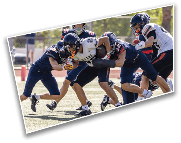 Several people wearing football helmets and uniforms are engaged in a tackle on a grassy field during a game. Description generated by AI