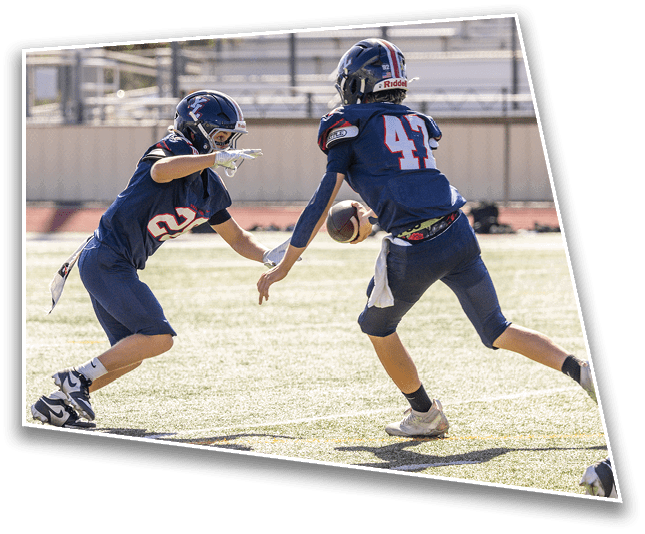 Two people in football uniforms and helmets are on a field with one handing off a football to the other. Description generated by AI