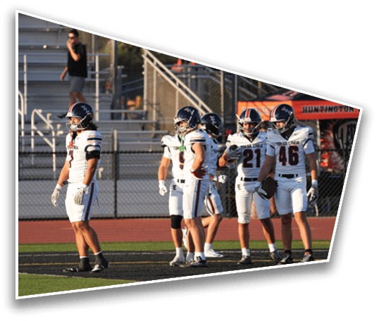 Several people in football uniforms and helmets stand on a field near bleachers and a tent in the background. Description generated by AI