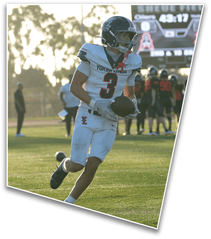 An athlete in a white football uniform with the number three is running on a grassy field holding a football while other players stand in the background. Description generated by AI