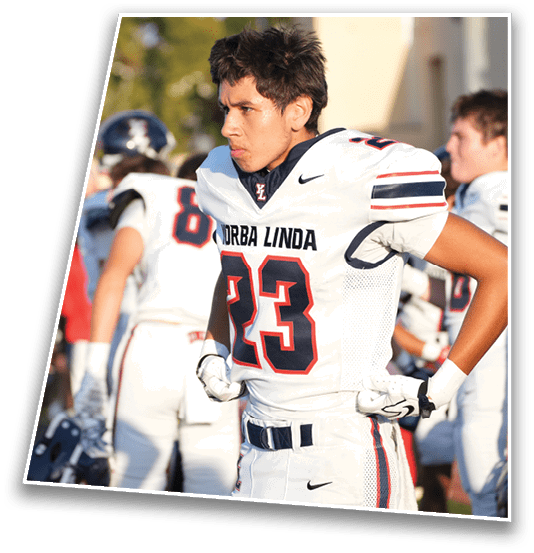 A person in a white football uniform with the number 23 and the words “DORBA LINDA" stands on the sidelines with hands on hips while other players and people are blurred in the background. Description generated by AI