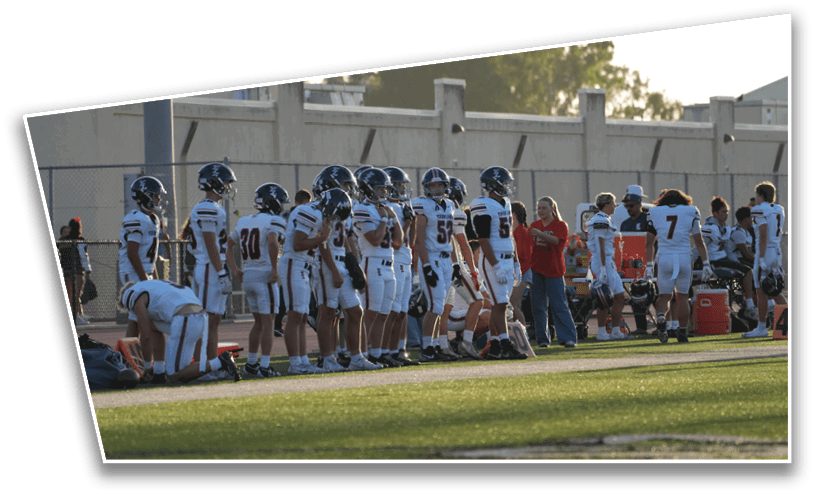 A group of people wearing football uniforms and helmets stand and kneel on the sidelines of a football field while others in casual clothing stand nearby. Description generated by AI