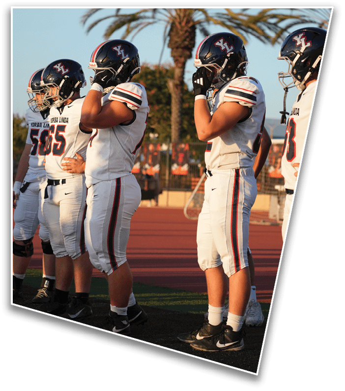 A group of people wearing white football uniforms and helmets stand in a line on a field with a palm tree and a track in the background. Description generated by AI