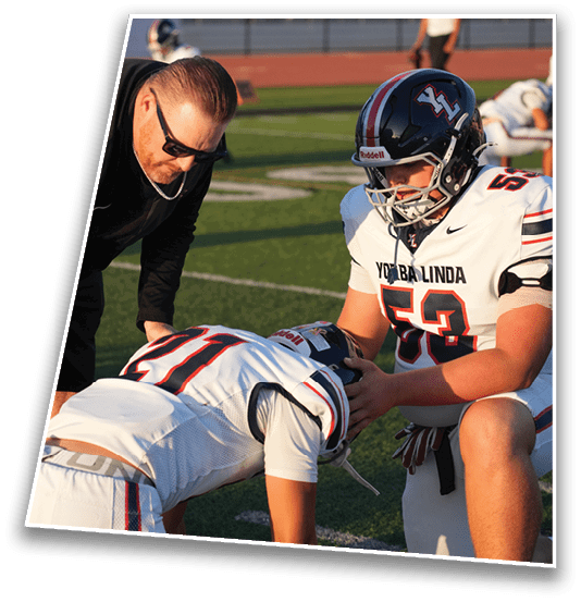 An adult wearing sunglasses is leaning down and talking to a person in a football helmet on a sports field with bleachers in the background. Description generated by AI