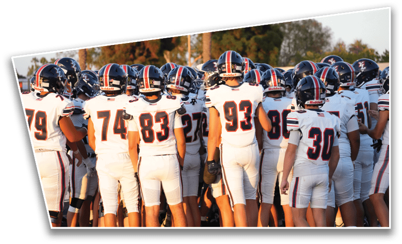 A group of people wearing white football uniforms and helmets are gathered closely together outdoors, likely preparing for a game or practice. Description generated by AI