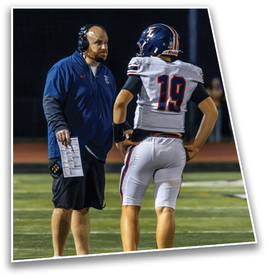 A person wearing a headset and holding a play sheet is talking to a person in a football uniform with the number 19 on the back on a sports field at night. Description generated by AI