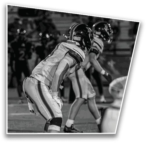 Two people in football uniforms and helmets are positioned on a field, appearing ready for action during a game. Description generated by AI