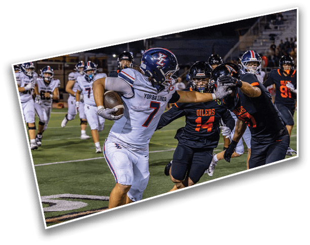 A person in a white football uniform with the number 7 is running with the ball while extending an arm to block two opposing players in black uniforms during a nighttime game on a football field. Description generated by AI
