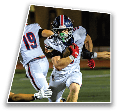 A person in a white football uniform with a helmet is running with the ball while being pursued by two other players on a football field at night. Description generated by AI
