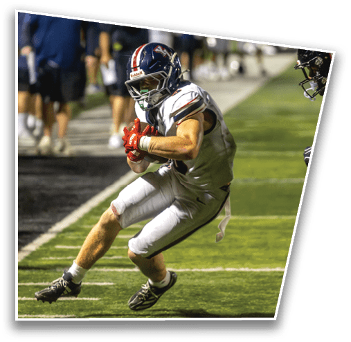 A person in a white football uniform is holding a football and crouching near the sideline while another person in a black football uniform approaches. Description generated by AI
