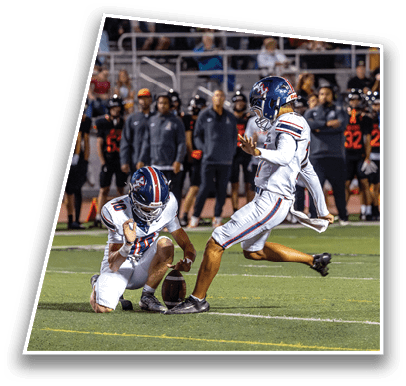 Two people in football uniforms are on a field with one holding the ball while the other prepares to kick it. Description generated by AI