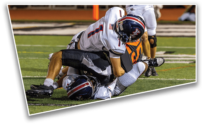 Two people in football uniforms are on the ground during a game on a field with one person wearing a white jersey and helmet and the other in a dark jersey. Description generated by AI