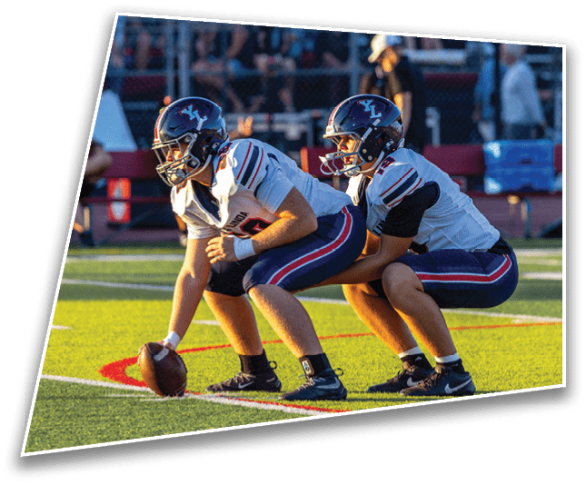 Two people in football uniforms and helmets are crouched on a field with one holding a football ready to snap it. Description generated by AI