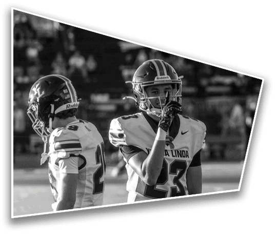Two people wearing football helmets and jerseys stand on a field, with one person holding a hand near their mouth as if signaling or speaking. Description generated by AI