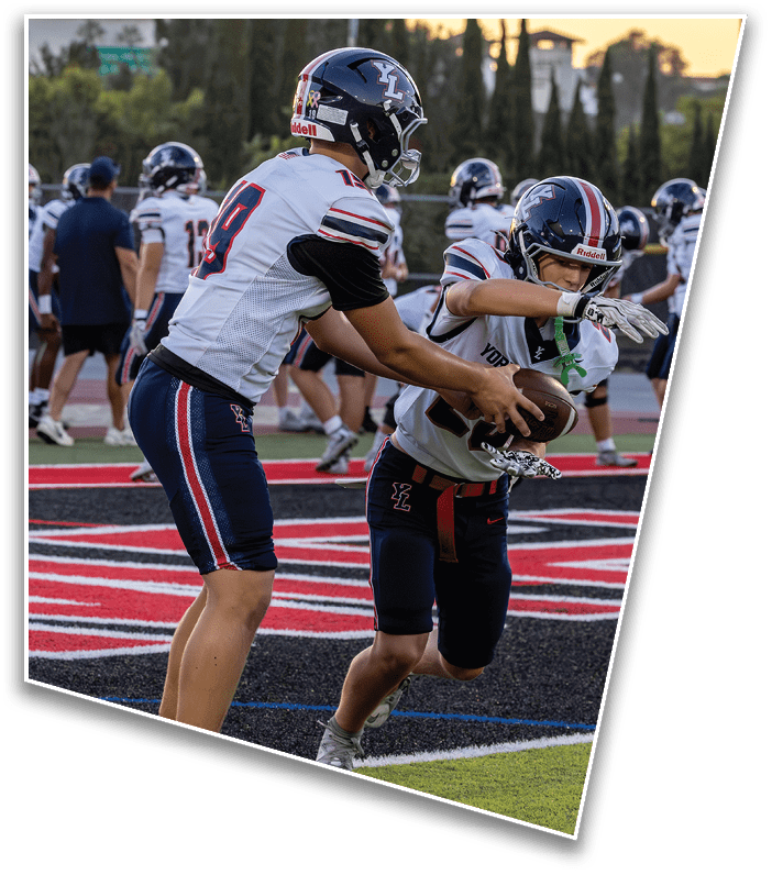 Two people in football uniforms are on a field with one handing off the ball to the other during a game or practice. Description generated by AI