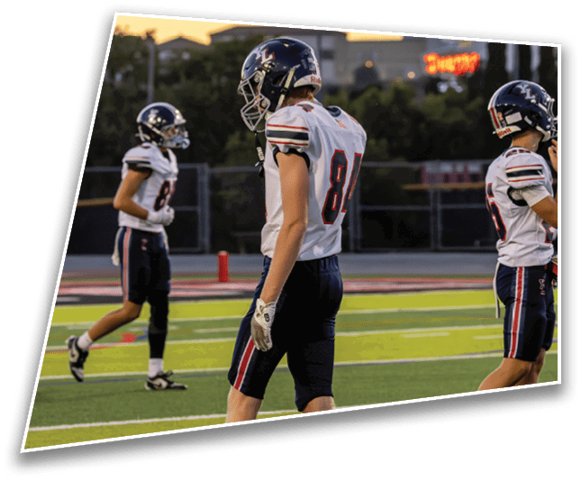 Three people wearing football uniforms and helmets stand on a field with yard lines visible during what appears to be a game or practice. Description generated by AI
