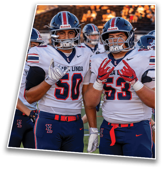 Two people in football uniforms with helmets stand on a field making hand gestures while others are visible in the background. Description generated by AI