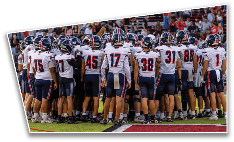 A group of people wearing football uniforms and helmets are huddled together on a field with spectators in the background. Description generated by AI