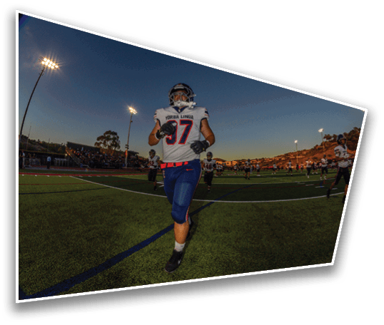 A person wearing a football uniform with the number 37 and the name Yorba Linda is running on a football field during dusk with other players and spectators in the background. Description generated by AI